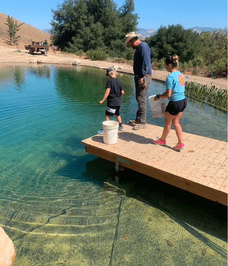 A man, a boy, and a young woman on a wooden dock at the edge of a clear, turquoise pond, possibly feeding fish, with dry hills and trees in the background.