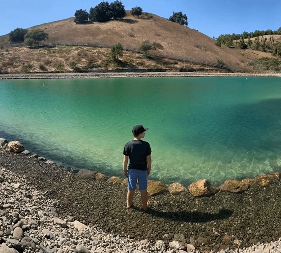 Person standing near a serene lake with hills in the background.